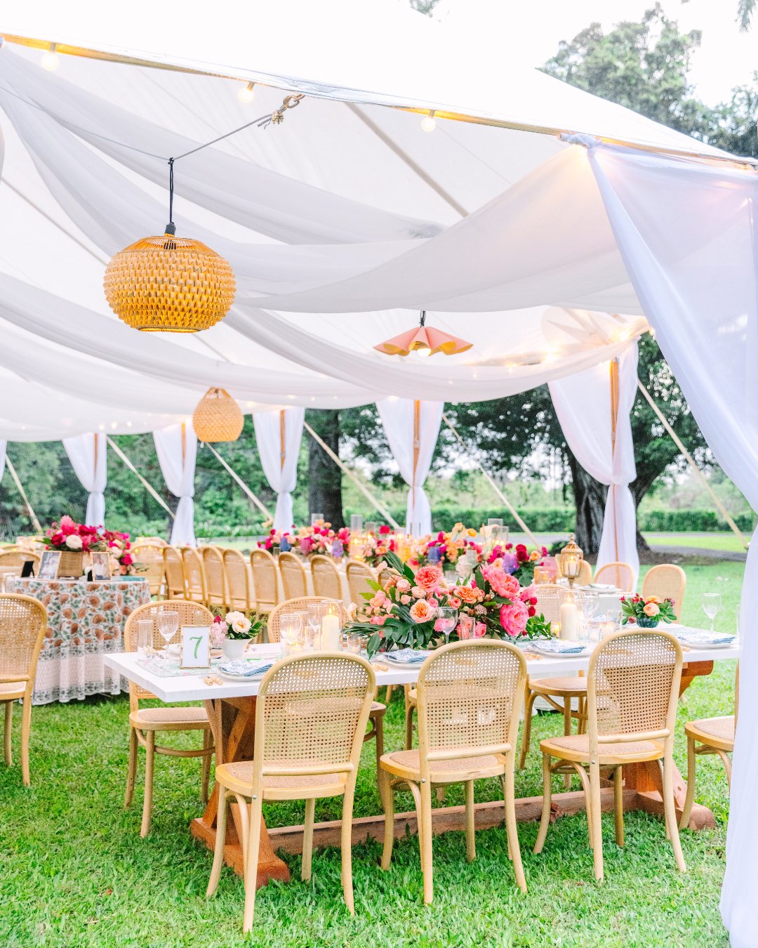 Outdoor event setup with wicker chairs around tables adorned with vibrant floral arrangements, under a white canopy with string lights and woven pendant lighting. Grassy area and trees visible.