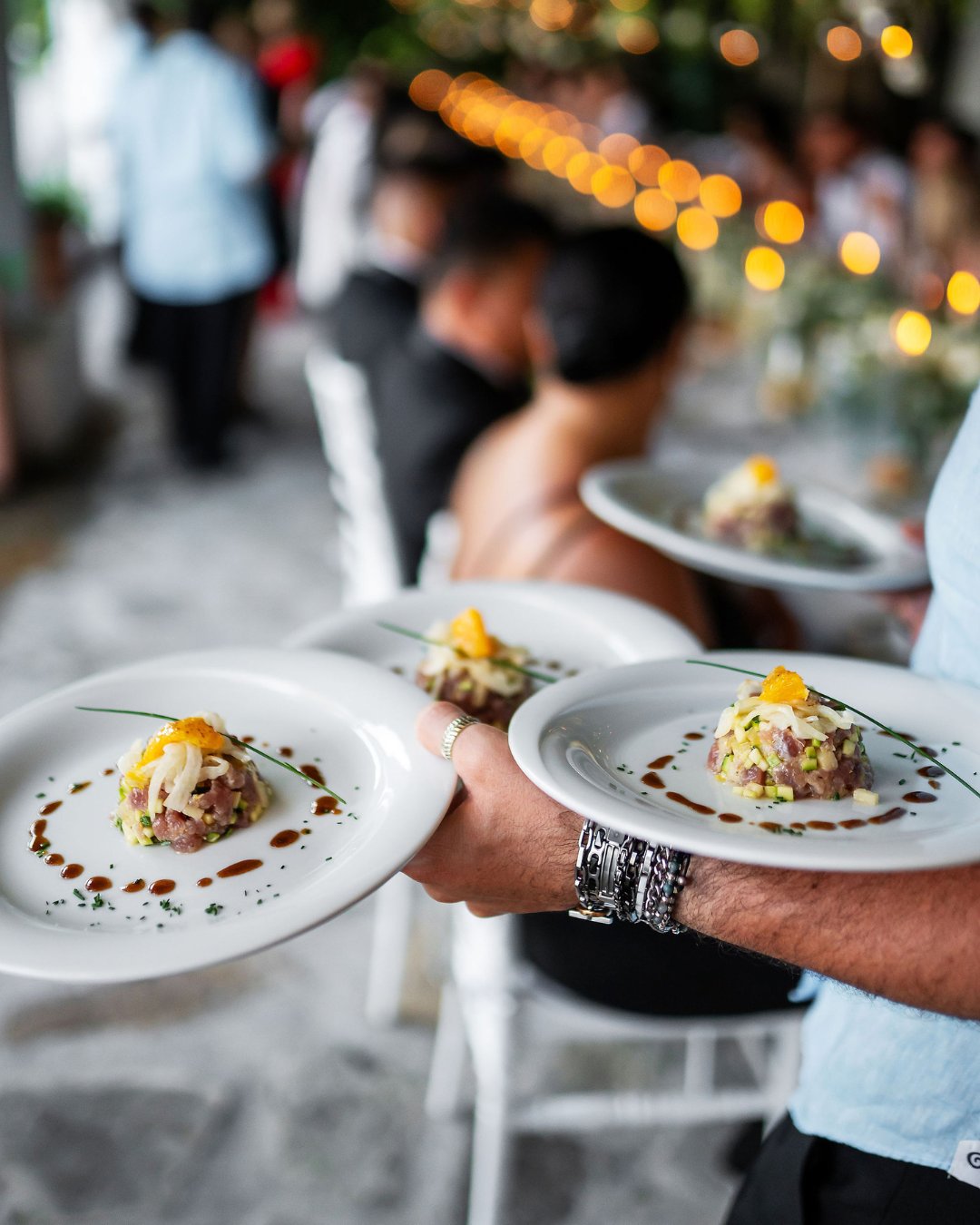 A person carries three plates of gourmet appetizers garnished with herbs and orange slices at an elegant dining event.