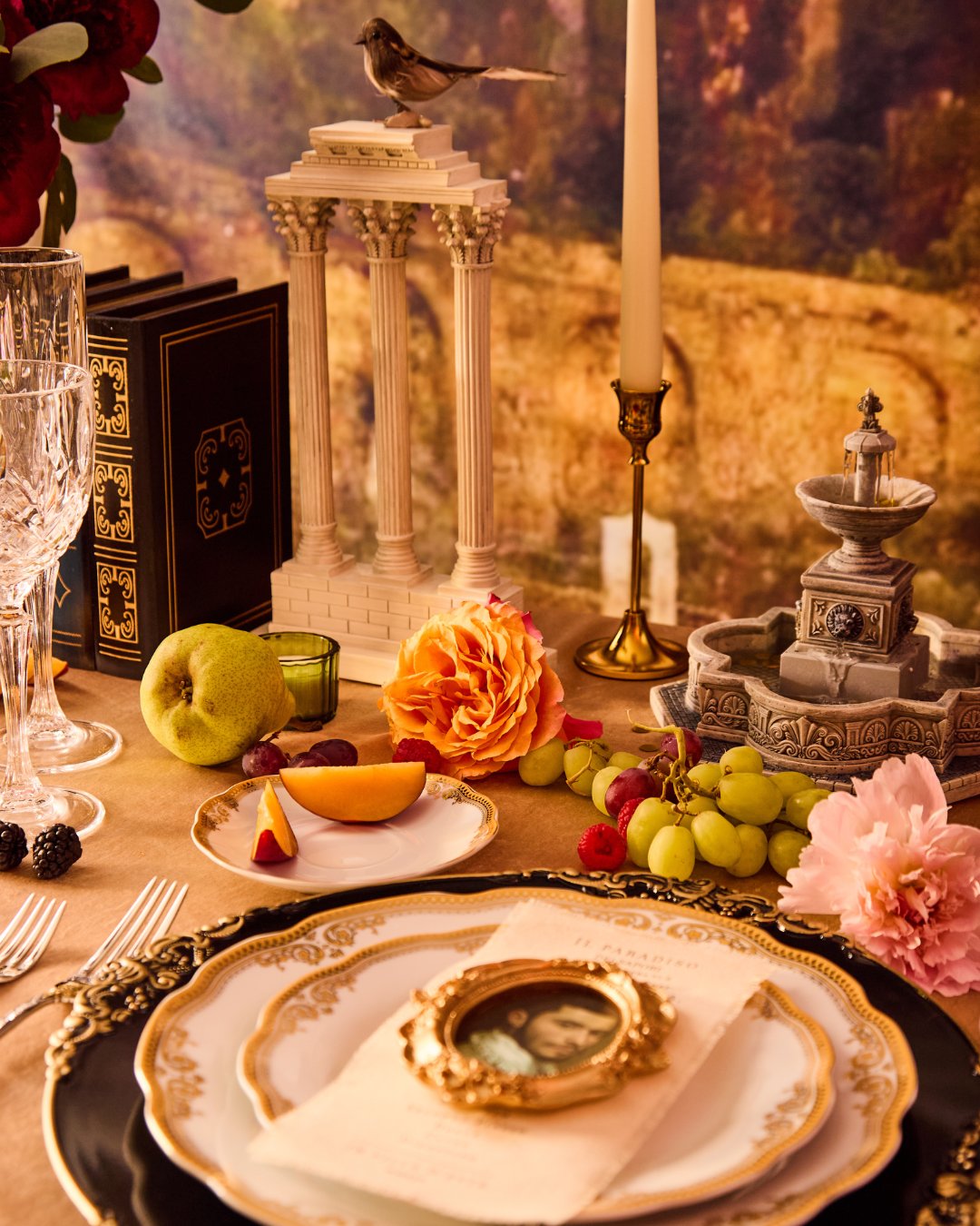 Elegant dinner table setting with decorative plates, fruit, flowers, crystal glassware, and a small statue. Background features a miniature column structure and a scenic mural.