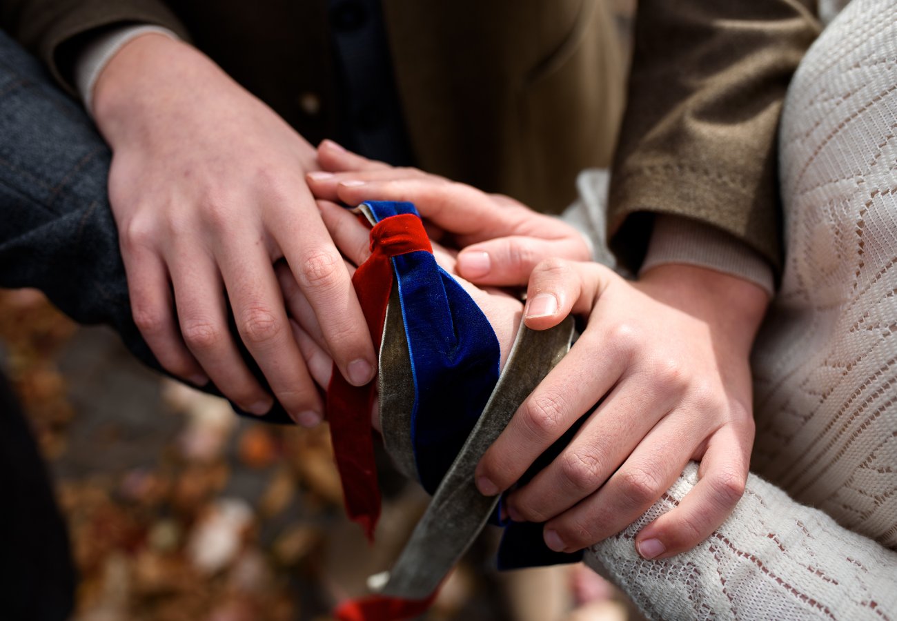 Four hands rest on top of each other, with one holding a blue and red velvet ribbon, suggesting a gesture of unity or support.
