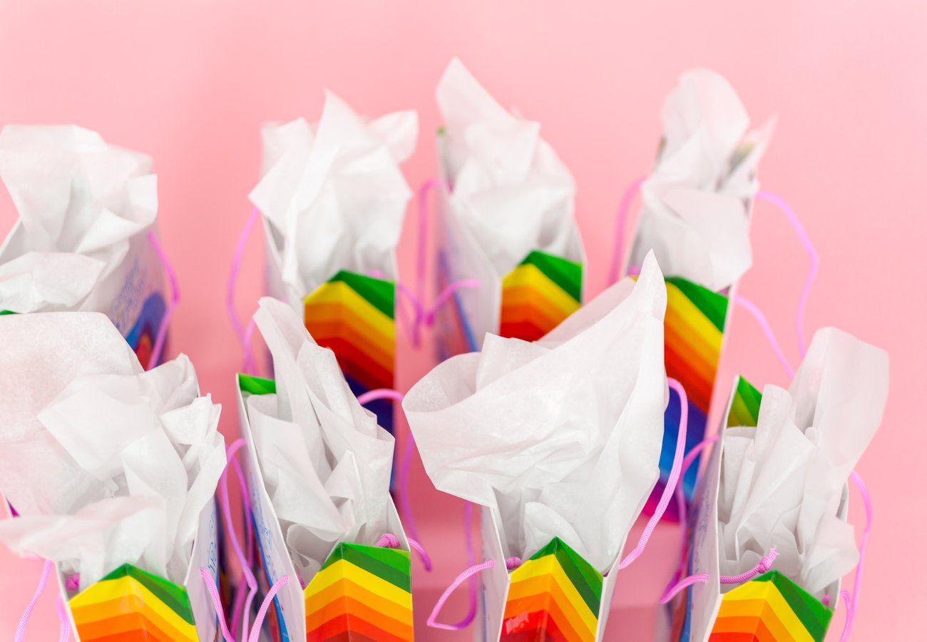 Several gift bags with rainbow-striped designs and white tissue paper are arranged on a pink background.