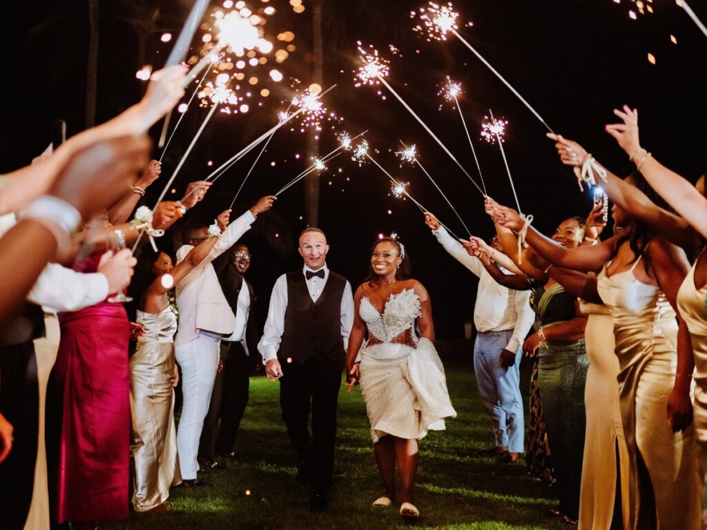 A bride and groom walk hand in hand under a tunnel of sparklers held by guests at an outdoor nighttime celebration.