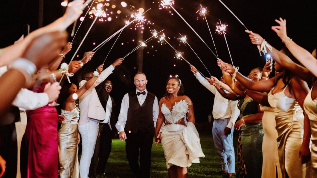 A bride and groom walk hand in hand under a tunnel of sparklers held by guests at an outdoor nighttime celebration.