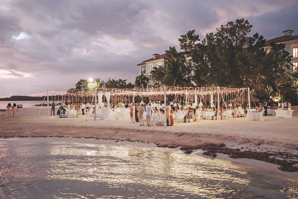 An outdoor event with string lights is set up on a sandy beach at sunset, with guests seated at round tables near a waterfront and trees in the background.