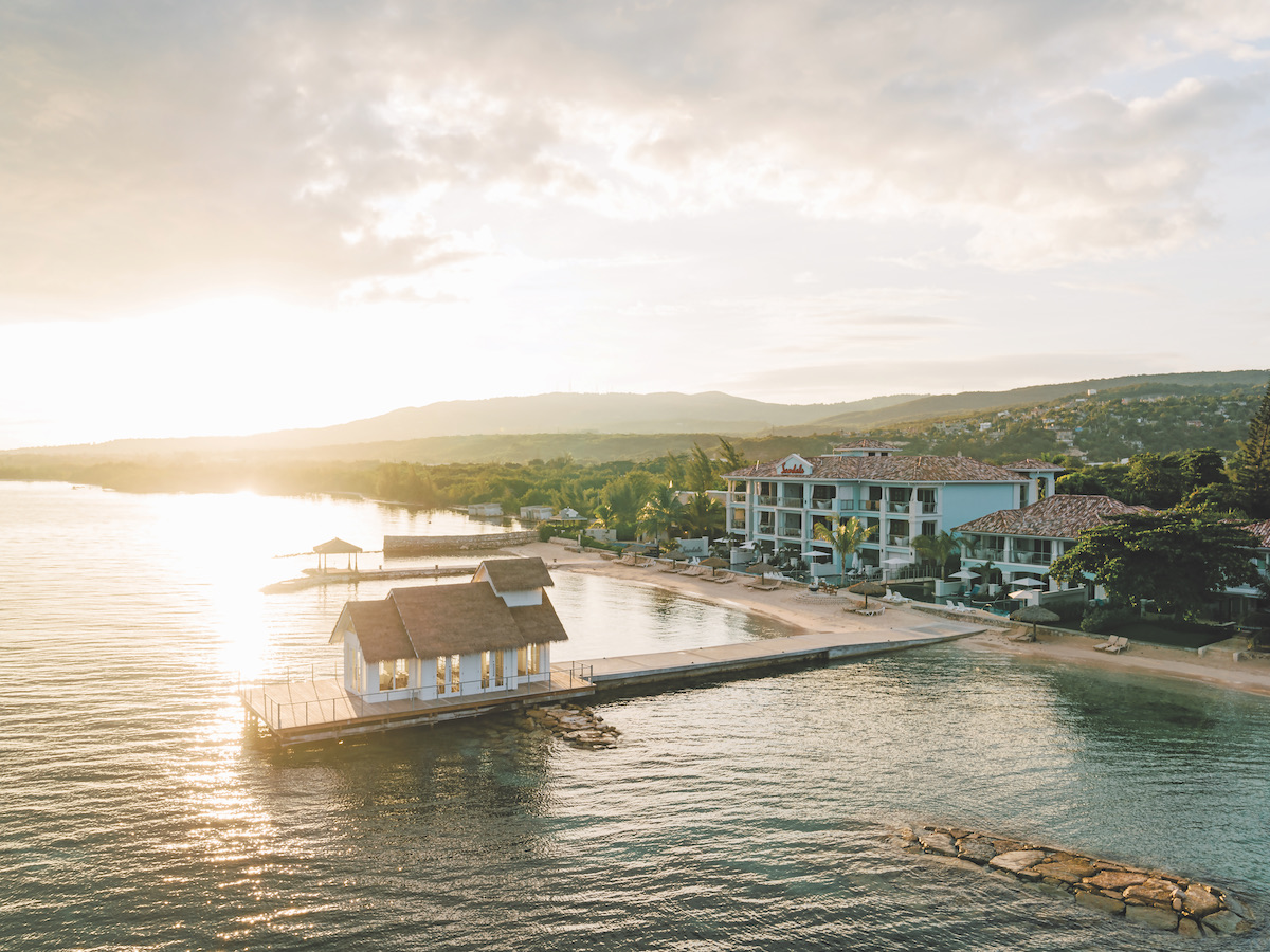A small pier with a white structure extends into calm water beside a beachfront resort at sunrise, with hills and greenery in the background.