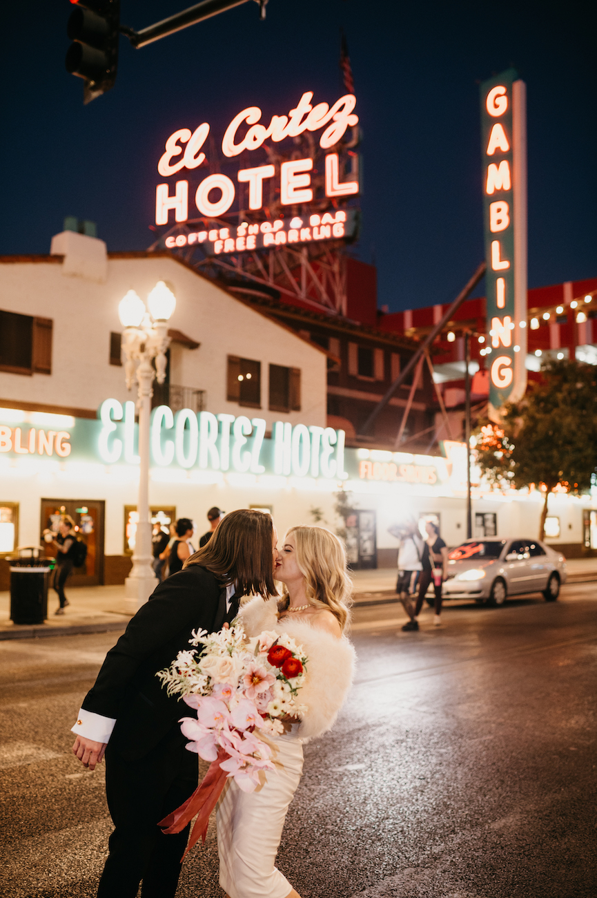 A couple dressed in wedding attire kisses on a street at night in front of the illuminated El Cortez Hotel and Gambling signs.