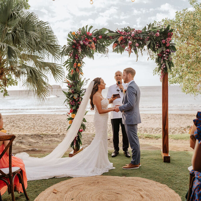 A bride and groom exchange vows under a floral arch at a beachside wedding ceremony, with guests seated nearby and the ocean in the background.