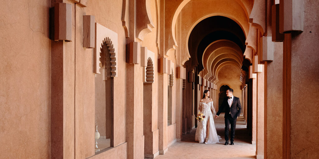 A couple dressed in formal wedding attire walks hand in hand through a corridor with arches and warm-toned walls.
