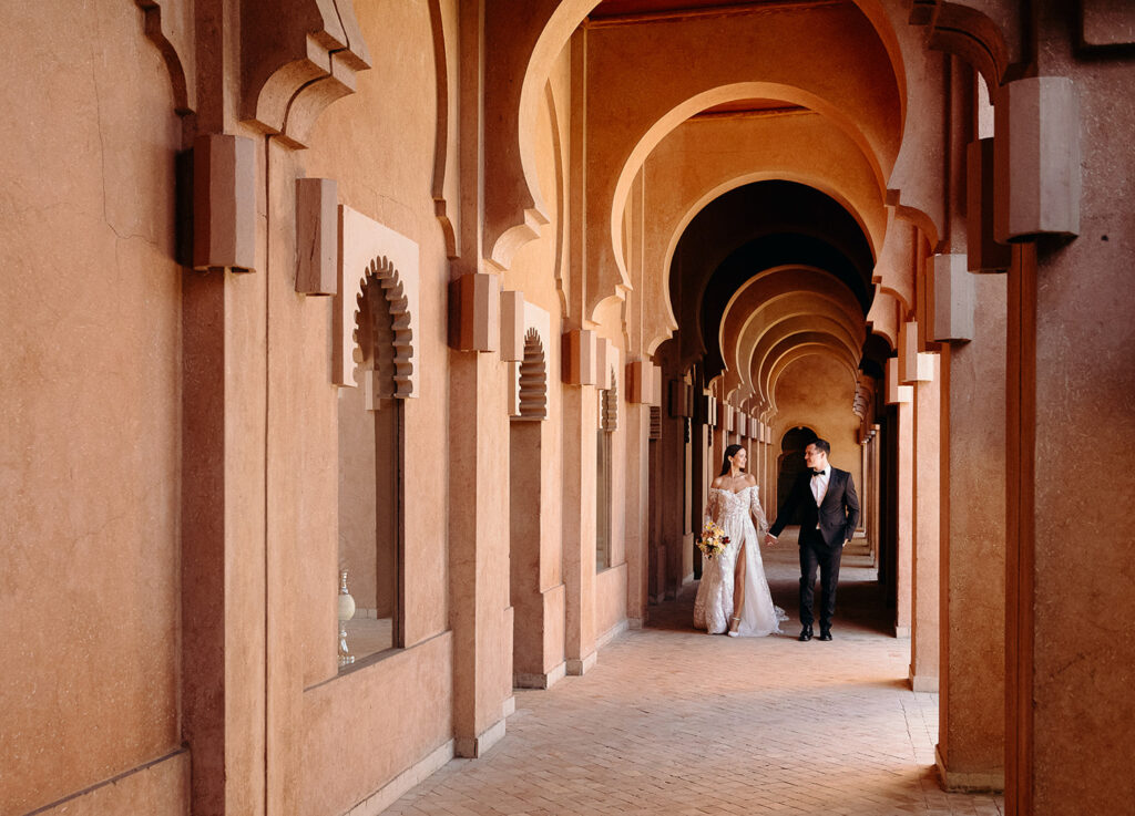 A couple dressed in formal wedding attire walks hand in hand through a corridor with arches and warm-toned walls.