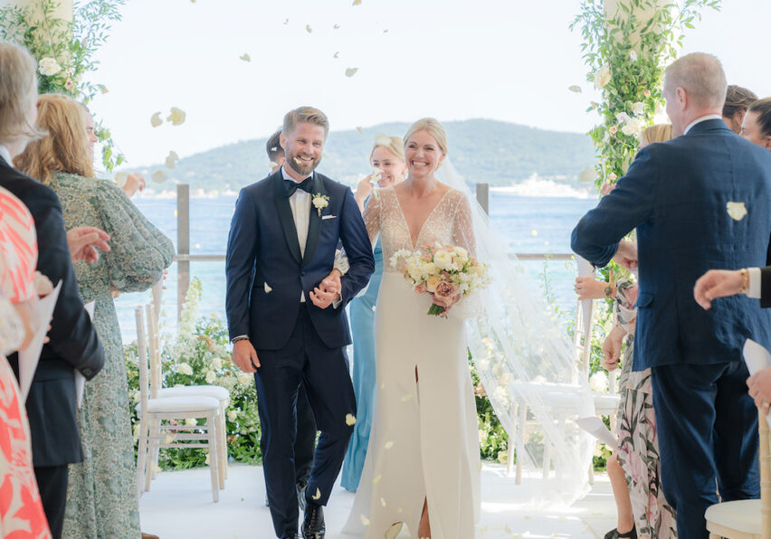 A bride and groom walk down the aisle outdoors, surrounded by guests throwing flower petals. The setting is decorated with greenery and overlooks a body of water.