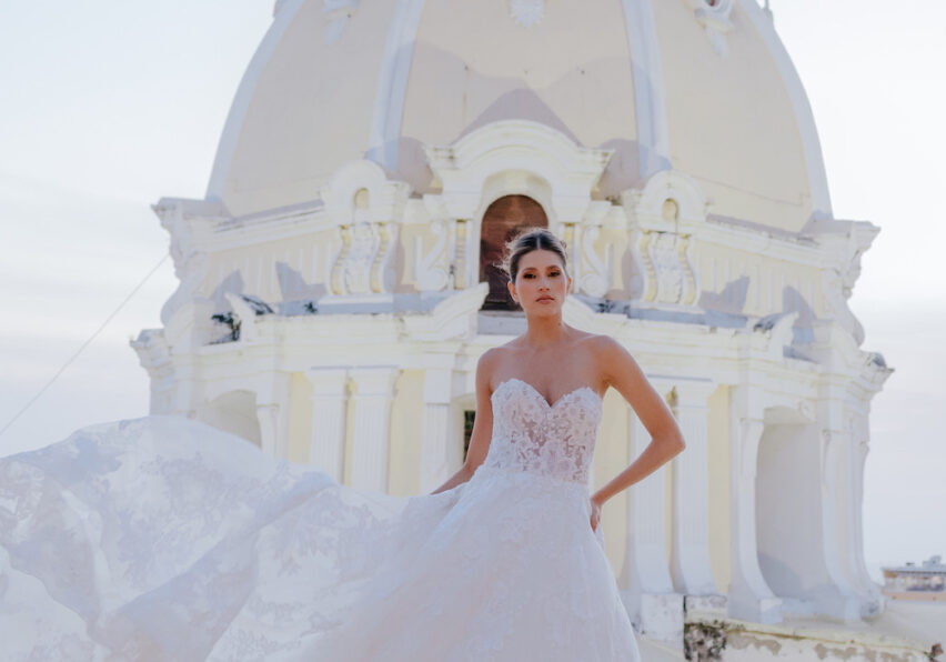 A woman in a strapless white wedding dress stands in front of a large white domed building, holding part of her dress out to the side.