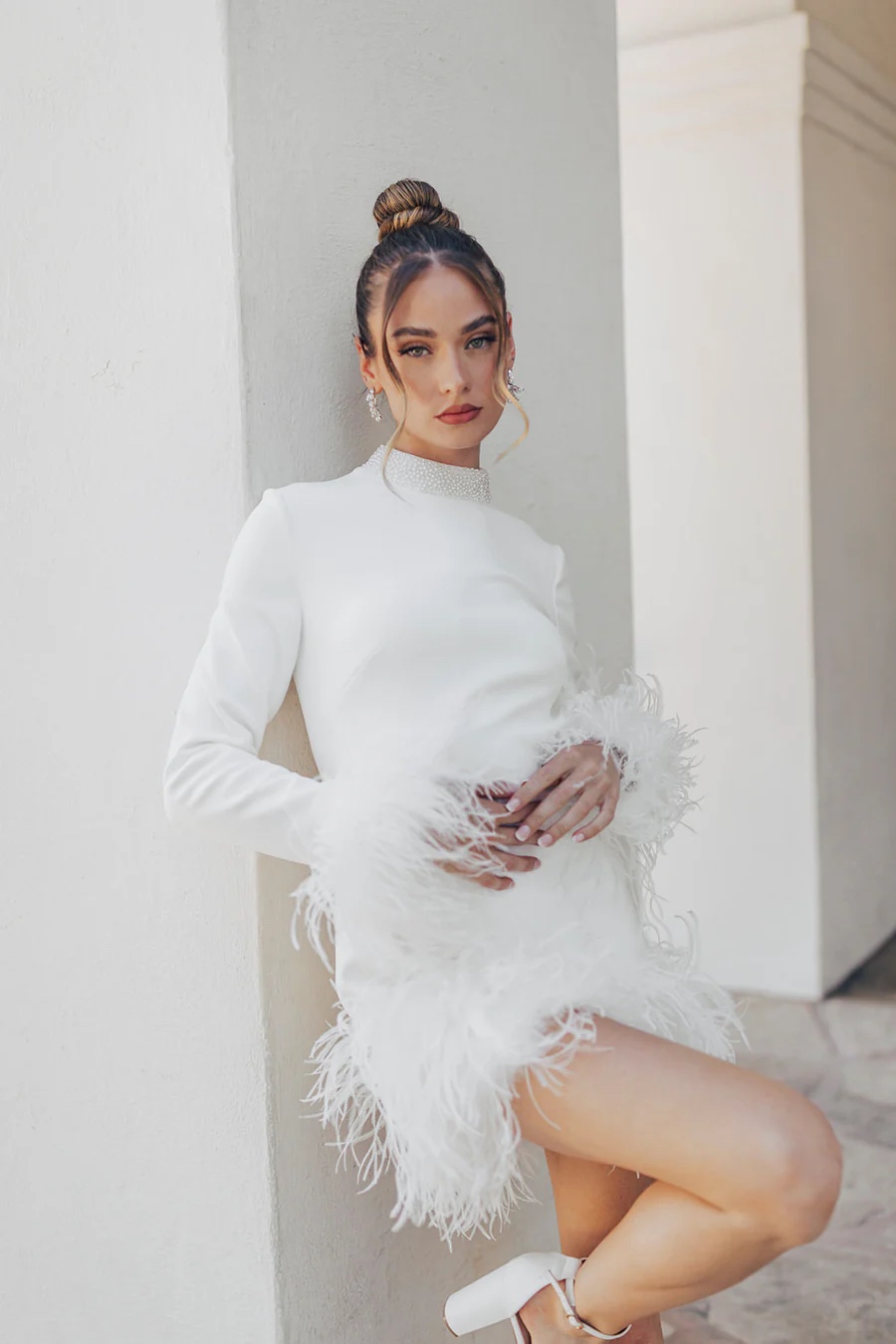 Woman leans against a white wall in a mini long sleeve crepe dress with faux feathers on the cuffs.