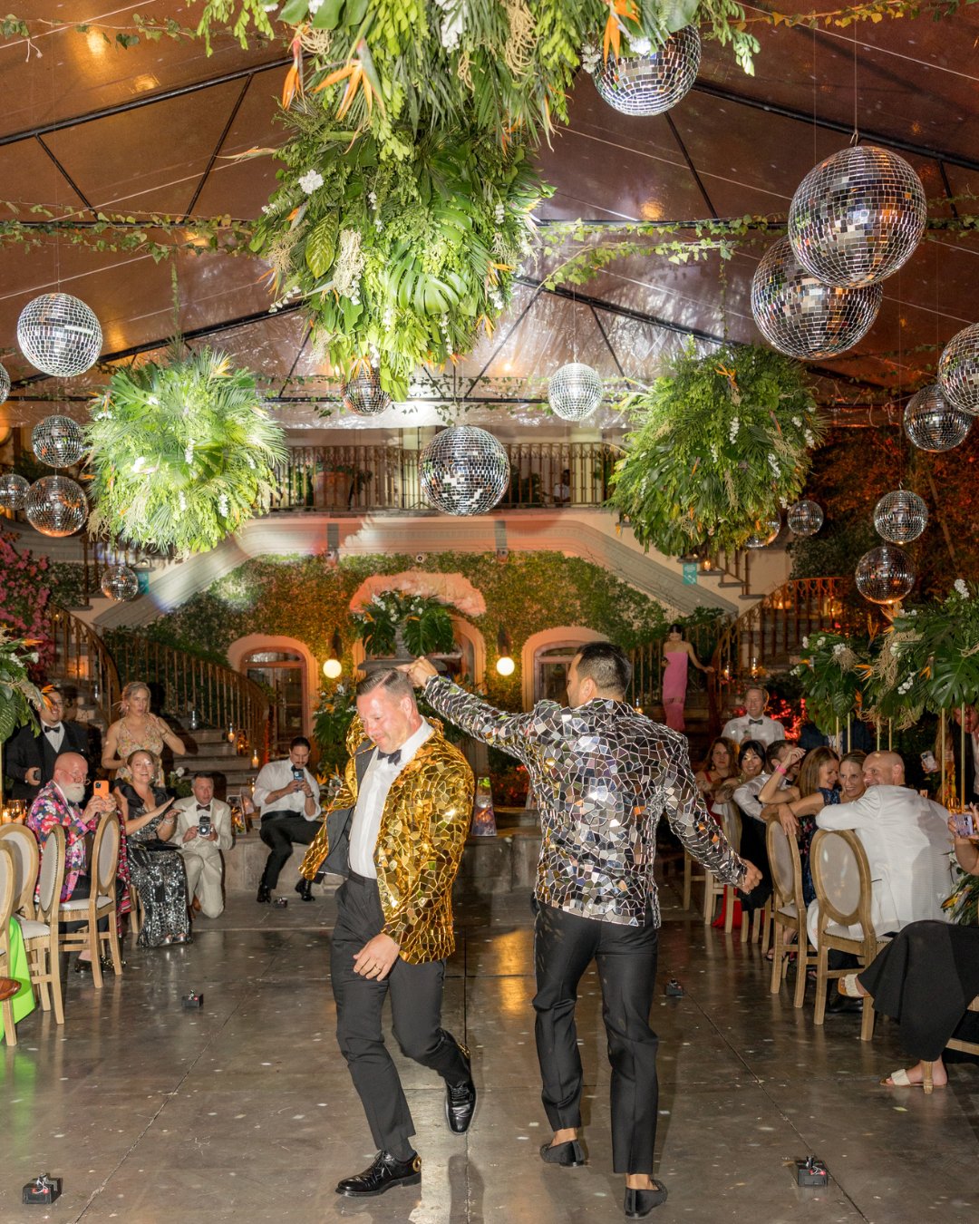 Two people in sequined jackets dance together under disco balls and greenery at a lively indoor event with seated guests watching.