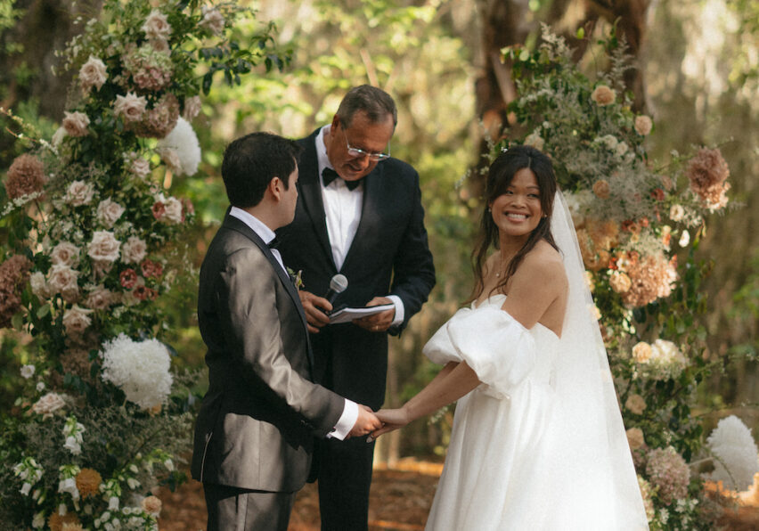 A bride and groom hold hands during an outdoor wedding ceremony, with a celebrant and wedding party standing nearby. Floral arrangements surround them.