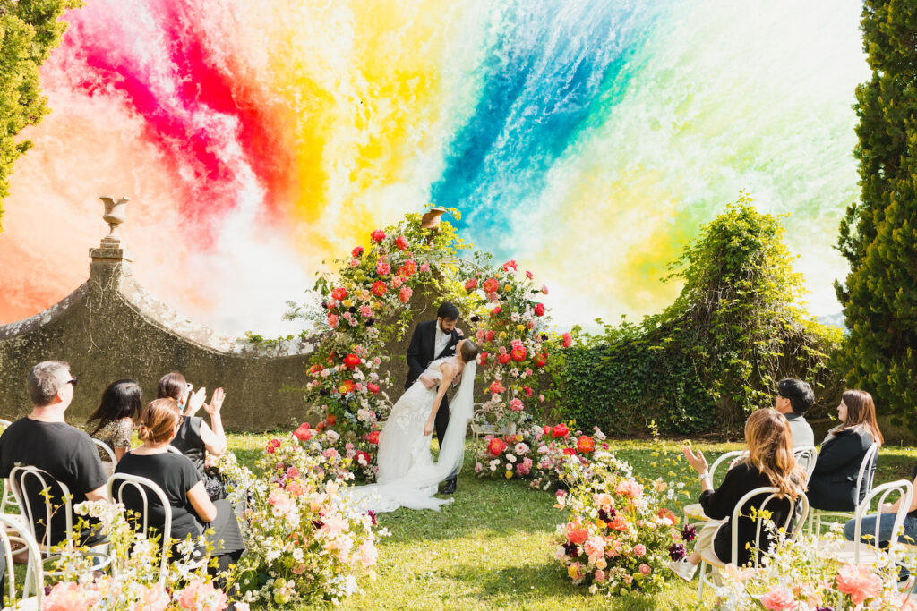 A bride and groom kiss under a floral arch at an outdoor wedding ceremony, with guests seated among flowers and a vibrant, colorful smoke fireworks in the background.