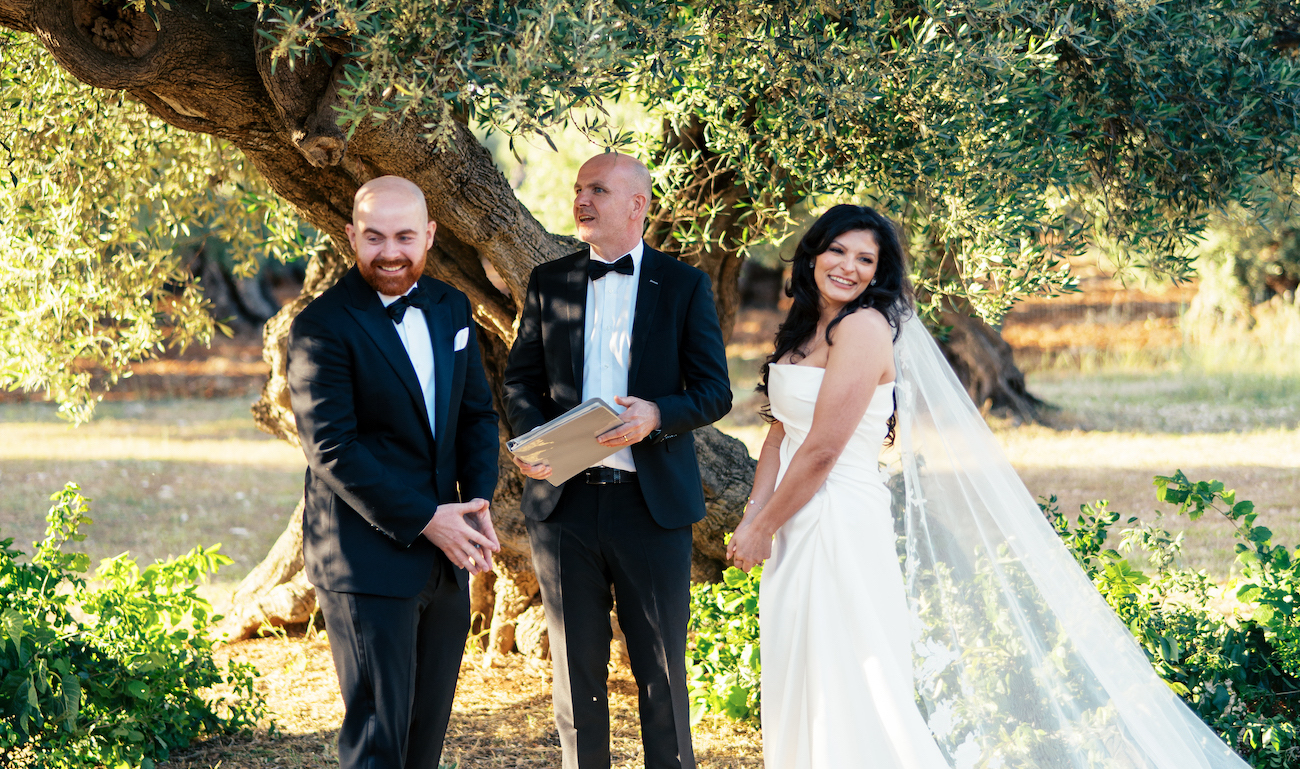 A bride and groom in formal attire stand with an officiant outdoors under a large tree during a wedding ceremony.