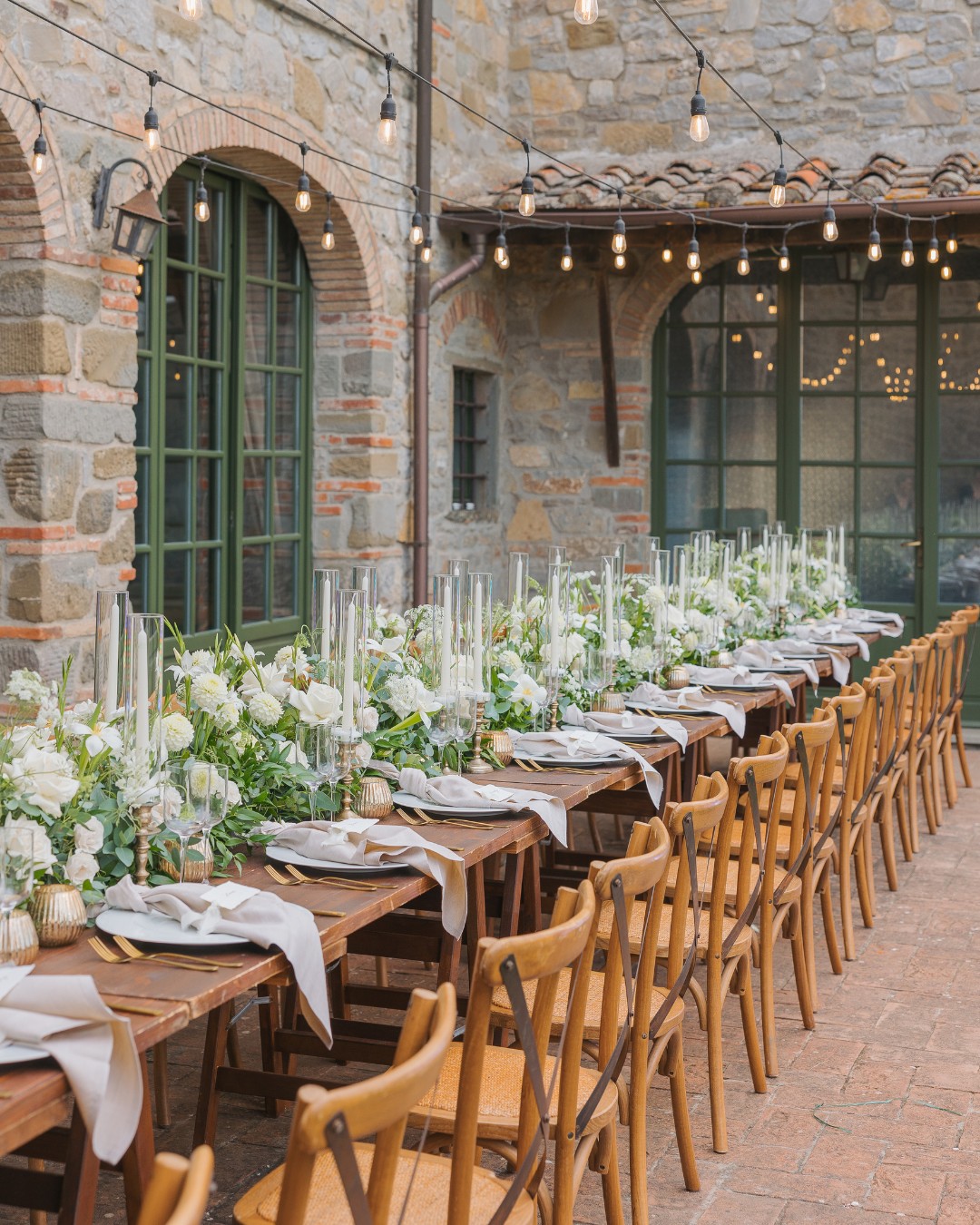 Long dining table set for an outdoor event, featuring white flowers, candles, and wooden chairs, against a stone building with string lights overhead.