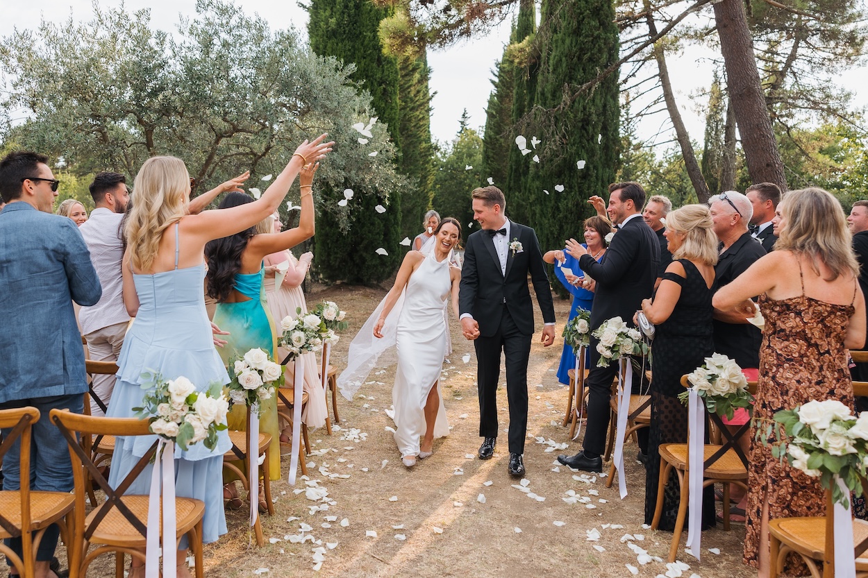 A bride and groom walk down the aisle outdoors as guests toss flower petals and applaud; white flowers decorate the aisle chairs.