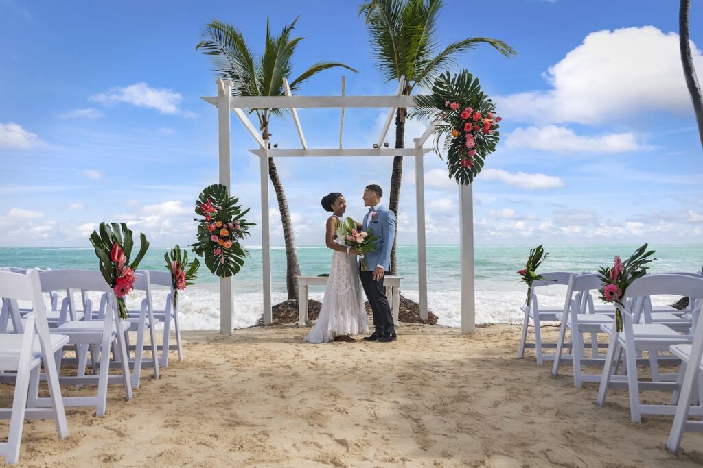 Bride and groom stand under a decorated white pergola on a sandy beach with palm trees, ocean, and flower arrangements in the background.