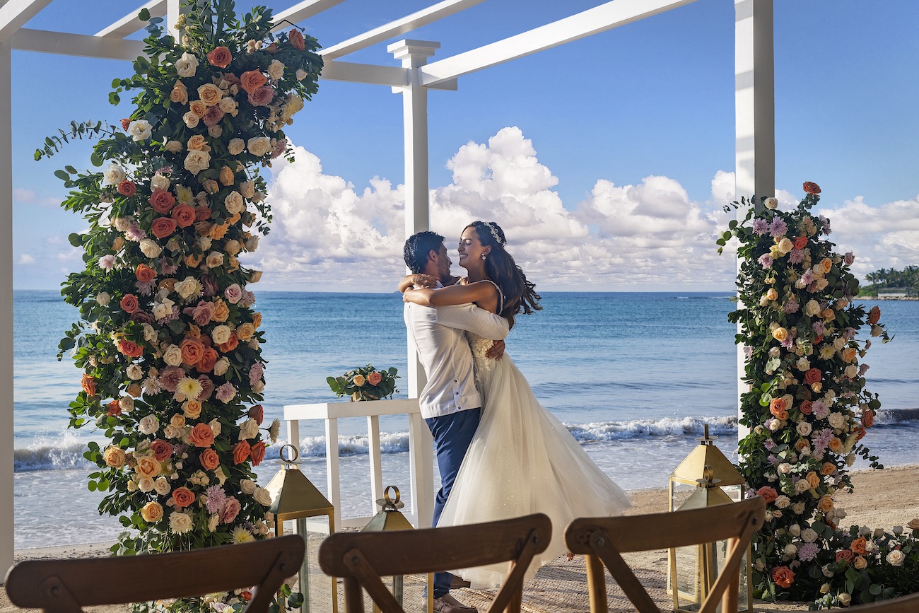 A bride and groom embrace under a floral arch at a beachside wedding ceremony with the ocean and clouds in the background.
