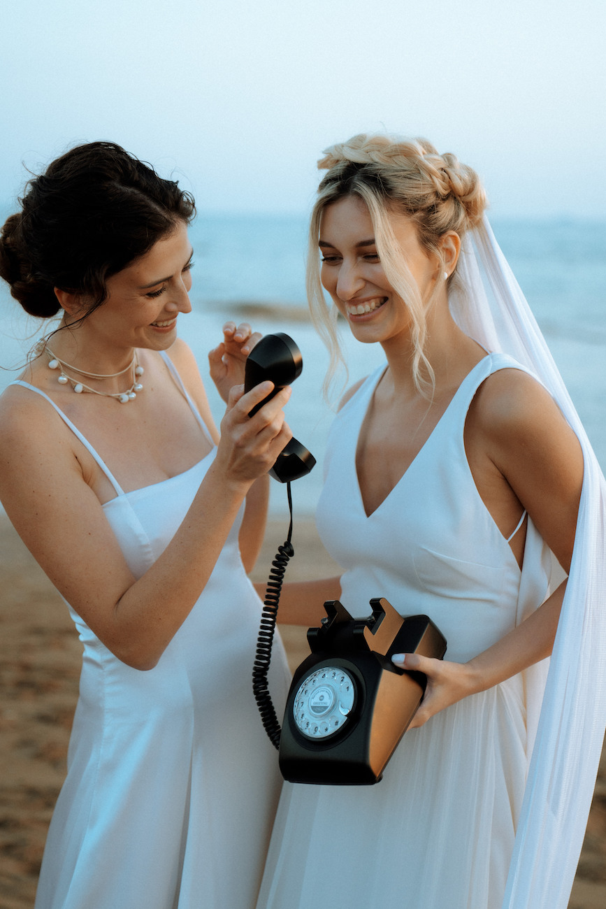 Two women in white dresses, one holding a retro rotary phone and smiling, standing together on a beach.