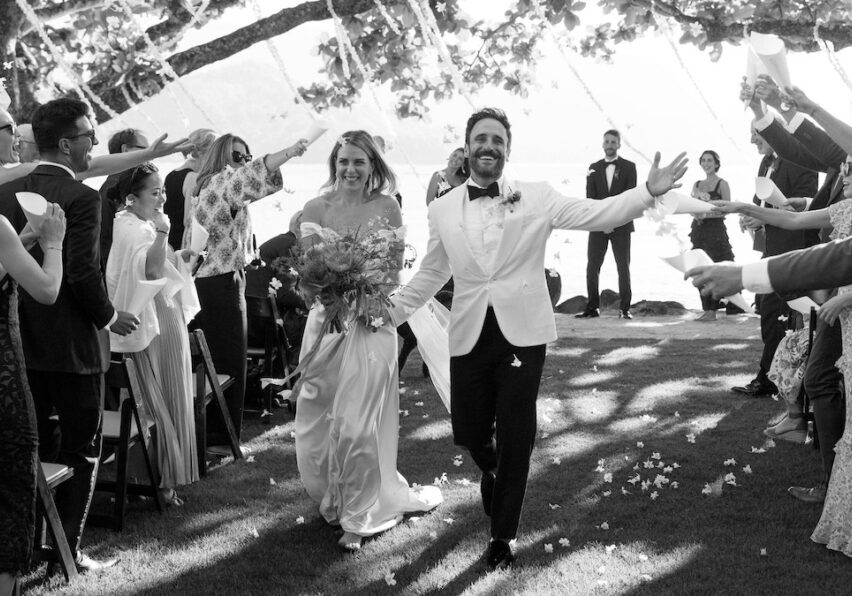 A newlywed couple walks down the aisle while guests cheer and toss petals. The event takes place outdoors under a tree canopy.