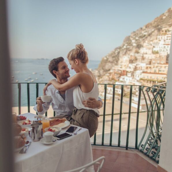 A couple sits closely together on a balcony overlooking a seaside town, having breakfast with food and drinks on the table.