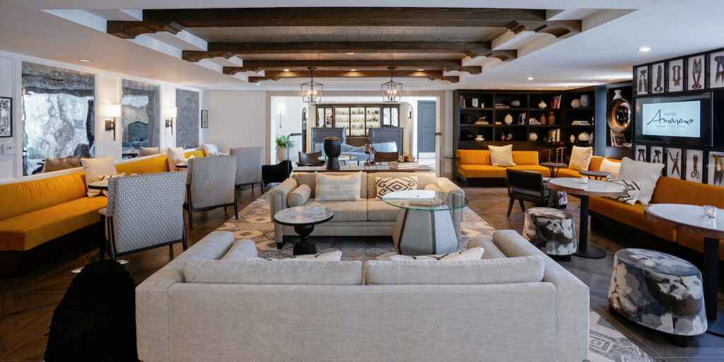 Spacious living room with beige sofas, armchairs, a glass coffee table, wooden ceiling beams, bookshelves, and a yellow couch.