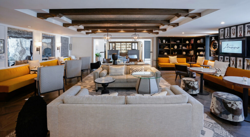 Spacious living room with beige sofas, armchairs, a glass coffee table, wooden ceiling beams, bookshelves, and a yellow couch.