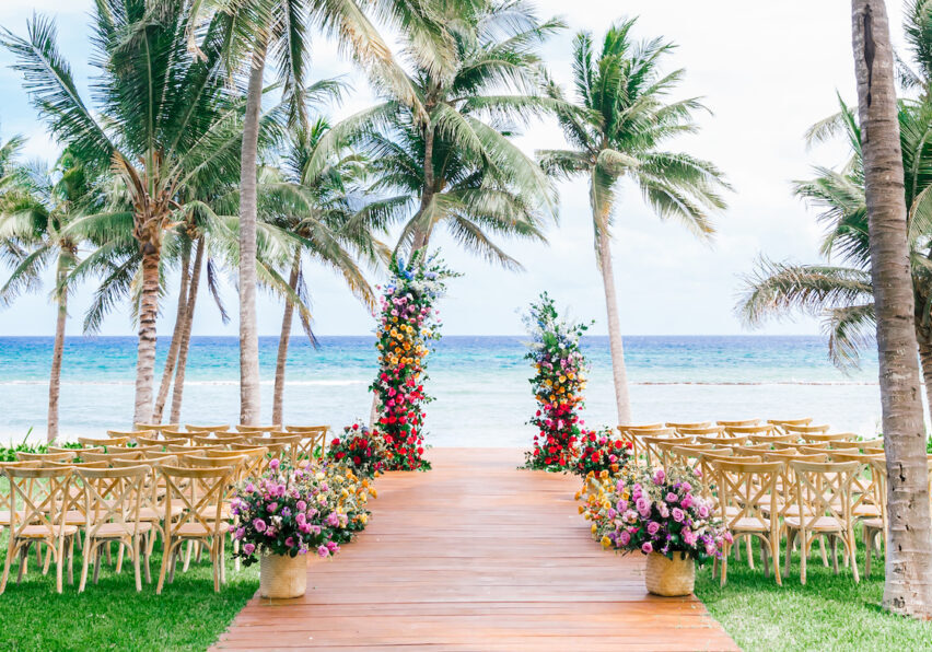A wooden aisle lined with floral arrangements and chairs leads to two colorful flower pillars on a beach, with palm trees and the ocean in the background.