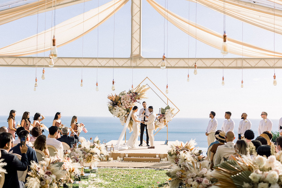 A couple stands at an altar adorned with flowers, exchanging vows under a canopy. Guests are seated on both sides, facing the ocean under a clear sky.