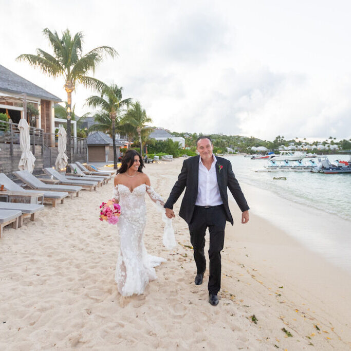 A bride and groom walk hand-in-hand along a sandy beach near lounge chairs and boats, with palm trees and a building in the background.