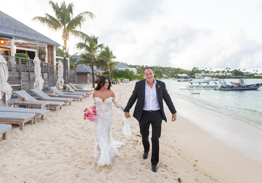 A bride and groom walk hand-in-hand along a sandy beach near lounge chairs and boats, with palm trees and a building in the background.
