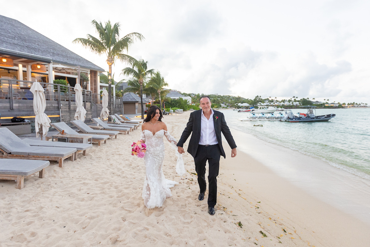 A bride and groom walk hand-in-hand along a sandy beach near lounge chairs and boats, with palm trees and a building in the background.