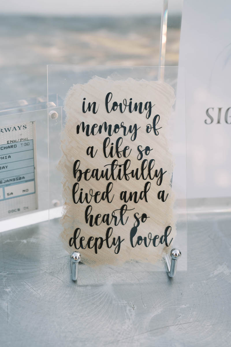 A memorial sign reads: "In loving memory of a life so beautifully lived and a heart so deeply loved," displayed on a clear stand.
