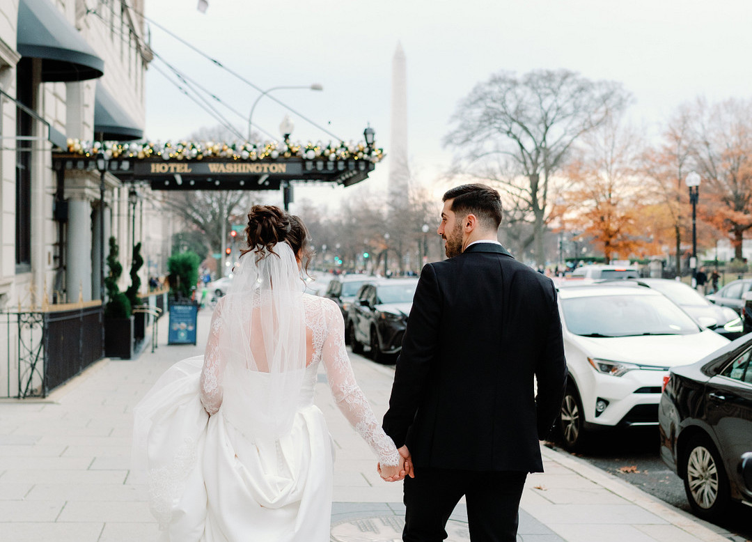 A bride and groom walk hand in hand on a city sidewalk. Trees and cars line the street, and a tall monument is visible in the background.