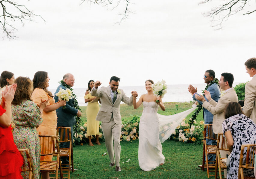 A bride and groom walk down an outdoor aisle, holding hands and smiling, as guests on both sides clap and cheer.