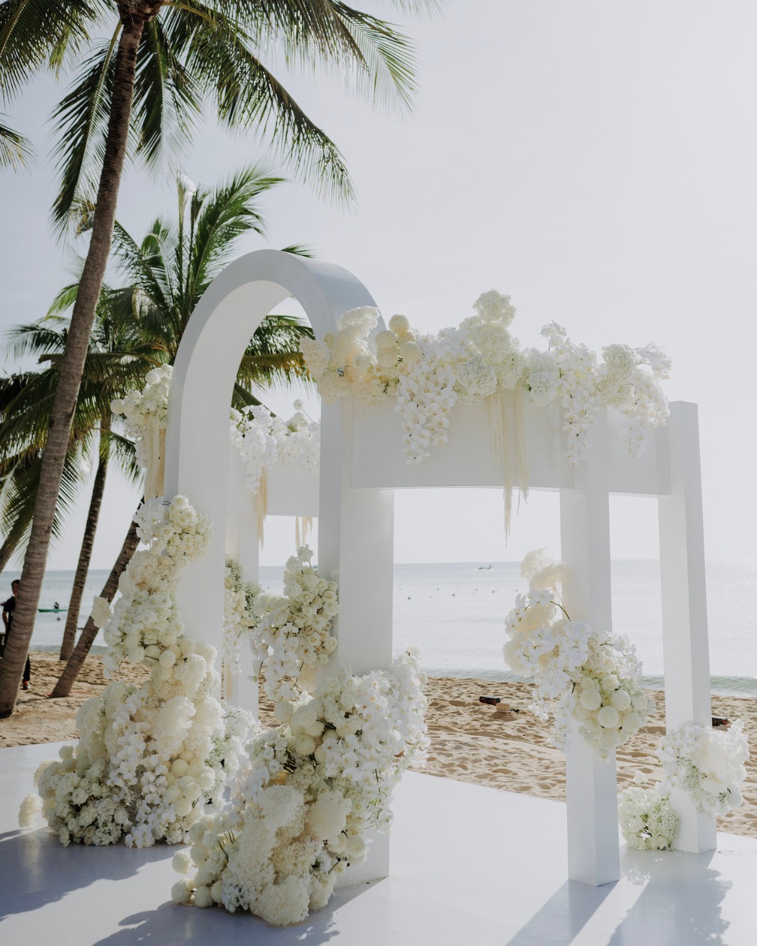 A beachside wedding arch decorated with white flowers, positioned on sand with palm trees and ocean in the background.