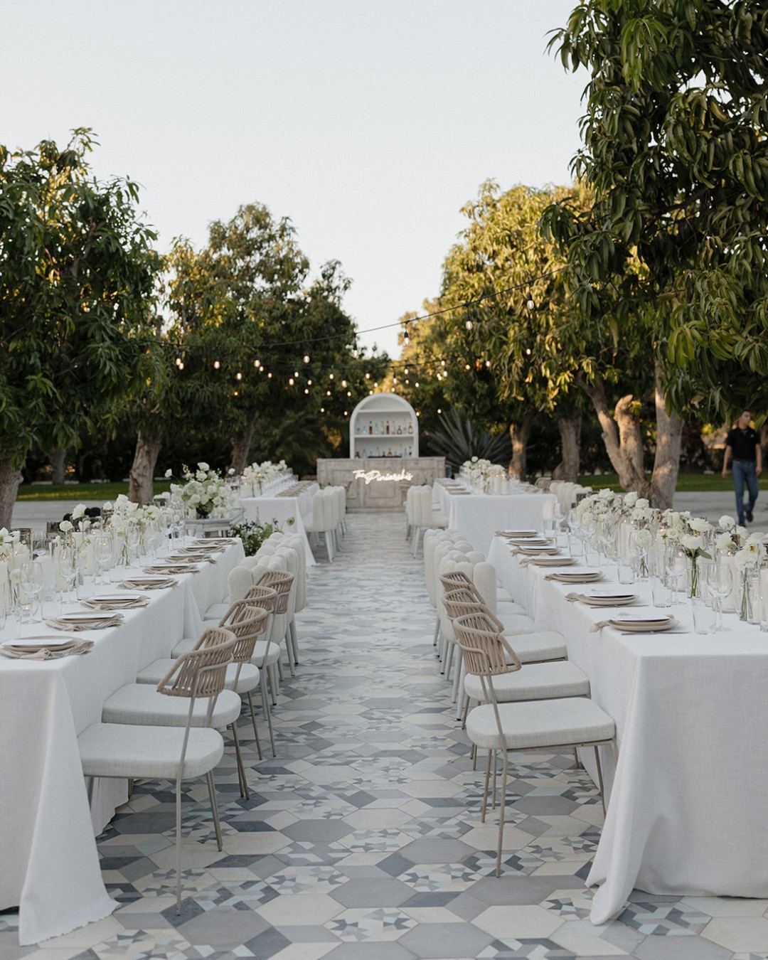 Outdoor dining setup with long tables covered in white tablecloths, surrounded by white chairs. Tables are adorned with glassware and white flowers. Overhead string lights add ambiance.