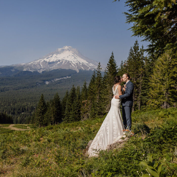 A bride and groom stand on grass surrounded by trees with a snow-capped Mt. Hood in the background under a clear sky.