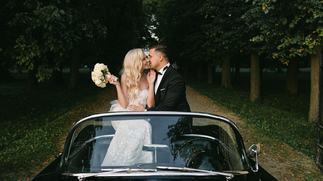 A bride and groom sit in a convertible on a tree-lined path. The bride holds a bouquet of white flowers, and both are dressed in formal attire, smiling at each other.