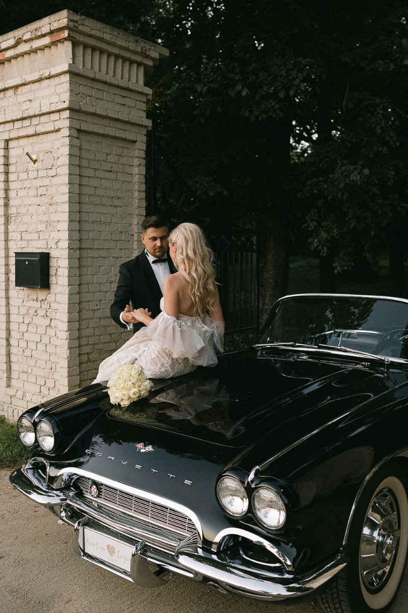 A couple in formal attire sit on a vintage black Corvette parked by a brick wall. The woman holds a bouquet of white roses.