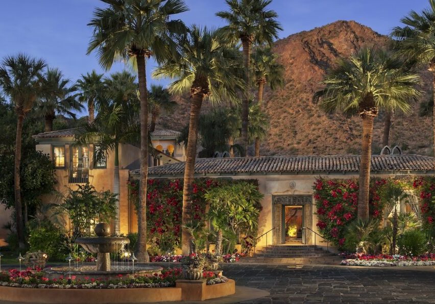 A Mediterranean-style villa with palm trees, flowering vines, and a courtyard fountain sits at the base of a rocky mountain at dusk.