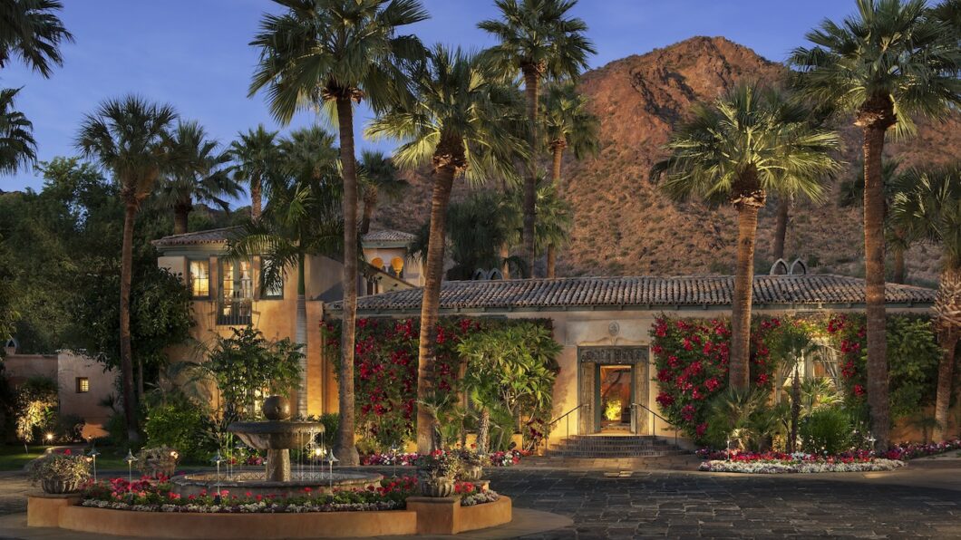 A Mediterranean-style villa with palm trees, flowering vines, and a courtyard fountain sits at the base of a rocky mountain at dusk.