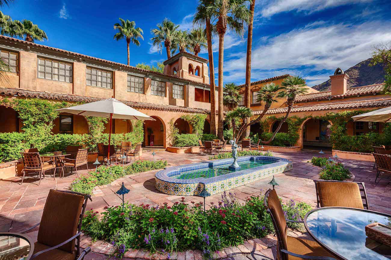 A sunny courtyard with a tiled fountain, surrounded by dining tables, lush plants, and a Spanish-style building with arches and palm trees.