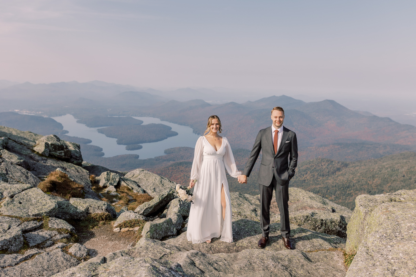 A bride and groom stand holding hands on a rocky mountain overlook with a river and mountains in the background.