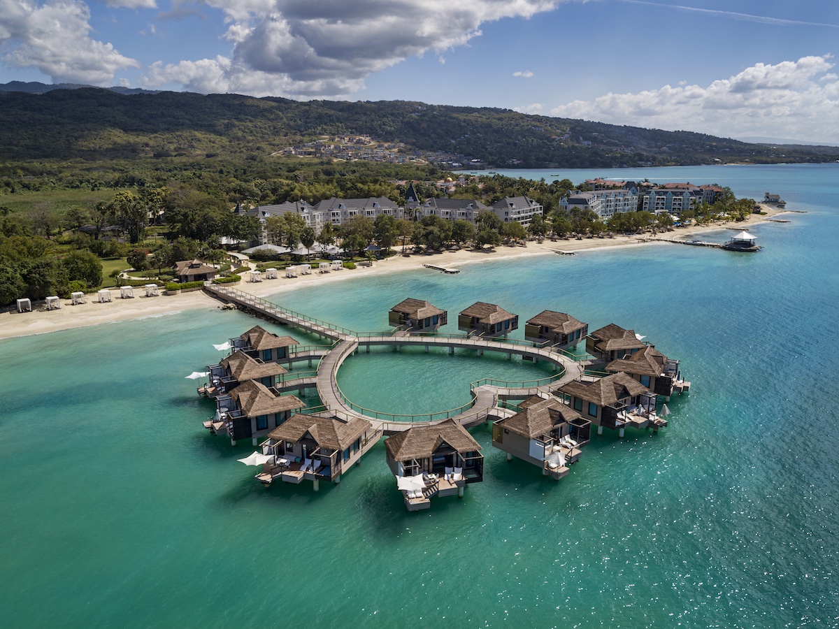 Aerial view of overwater bungalows arranged in a circle near a sandy beach, with a resort complex and green hills in the background.