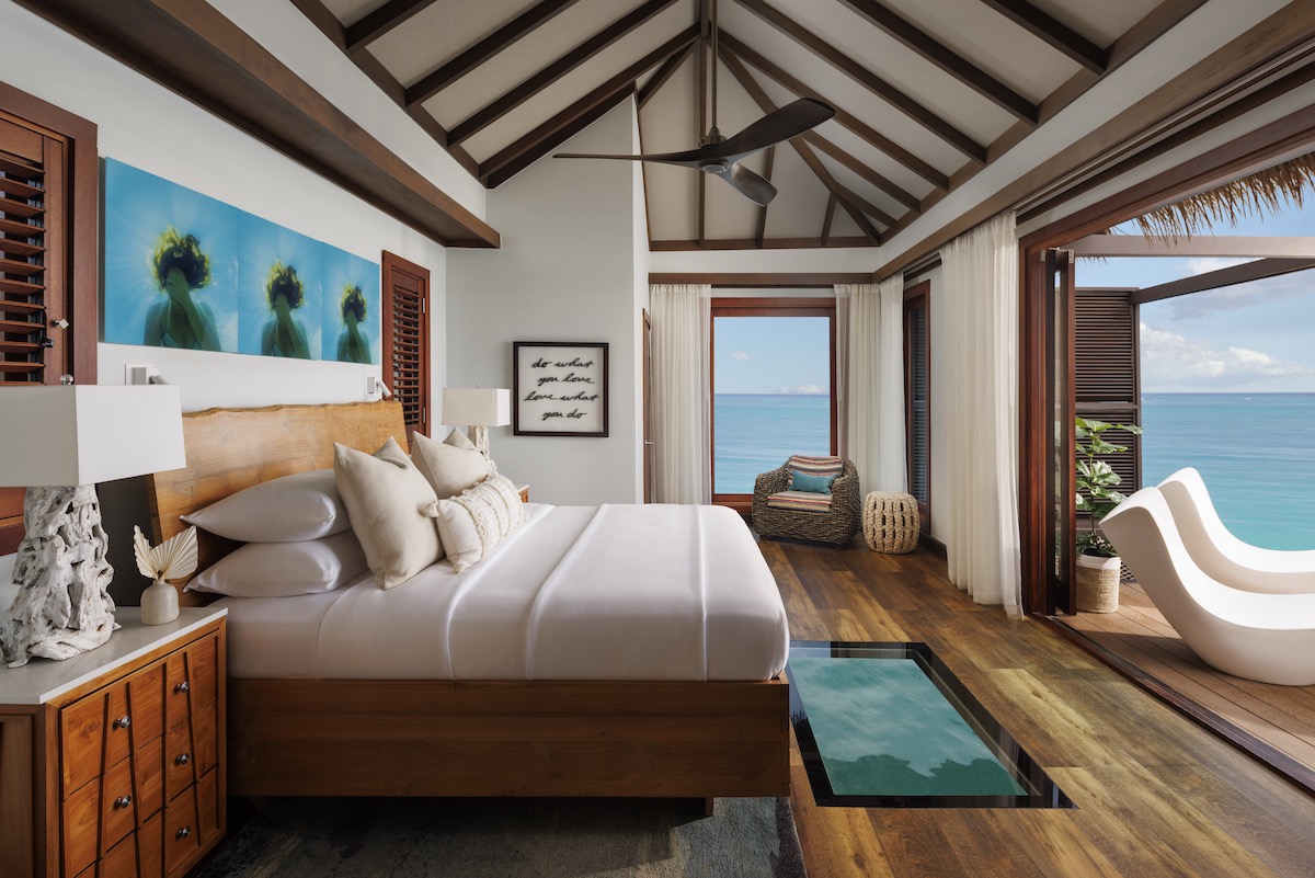 Modern tropical bedroom with wood accents, large bed, wall art, and sliding doors opening to an ocean view with balcony seating; glass panel in floor shows water below.