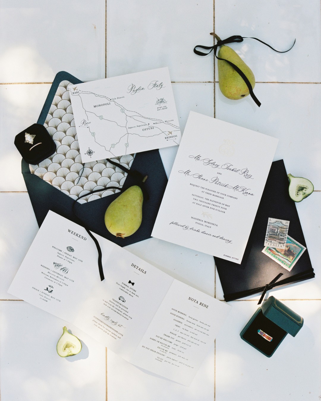 Flat lay of wedding invitation suite with envelopes, a map, pears, rings in boxes, vintage stamps, and a weekend itinerary, arranged on a white tiled surface.
