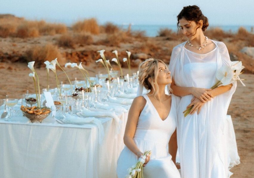 Two women in white dresses pose beside an outdoor table set with white flowers and tableware on a beach, with dry grass and the sea in the background.